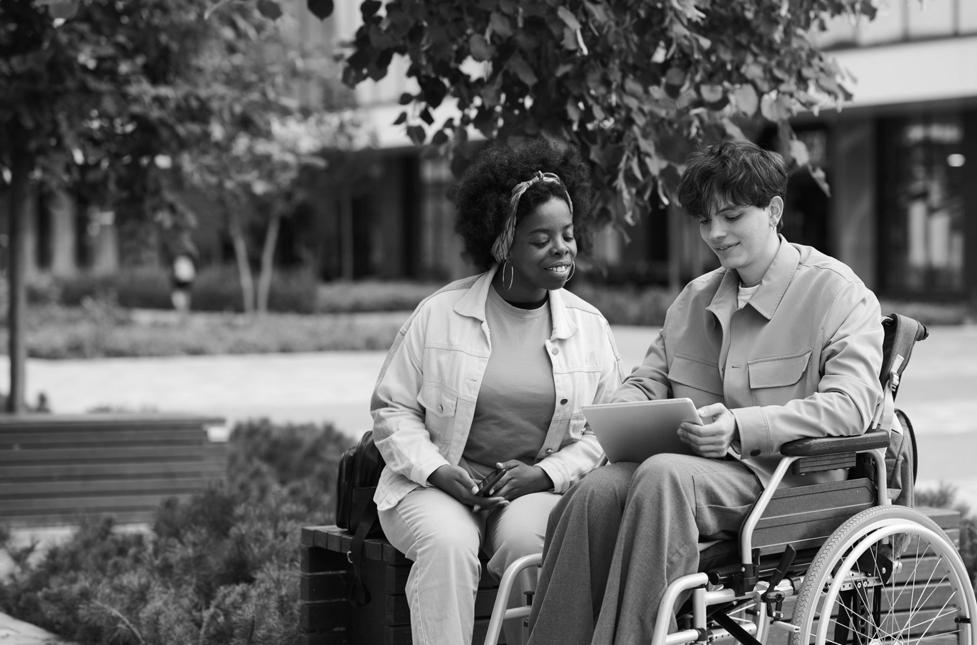 two people sit together in a park, as one in a wheelchair shows the other something on a computer