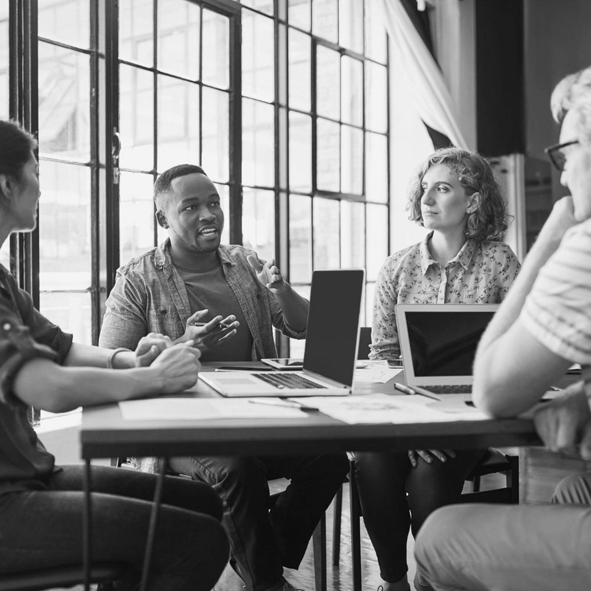 a diverse group of people sit at a table with computers in front of them as one man speaks and gestures with his hands and the others listen