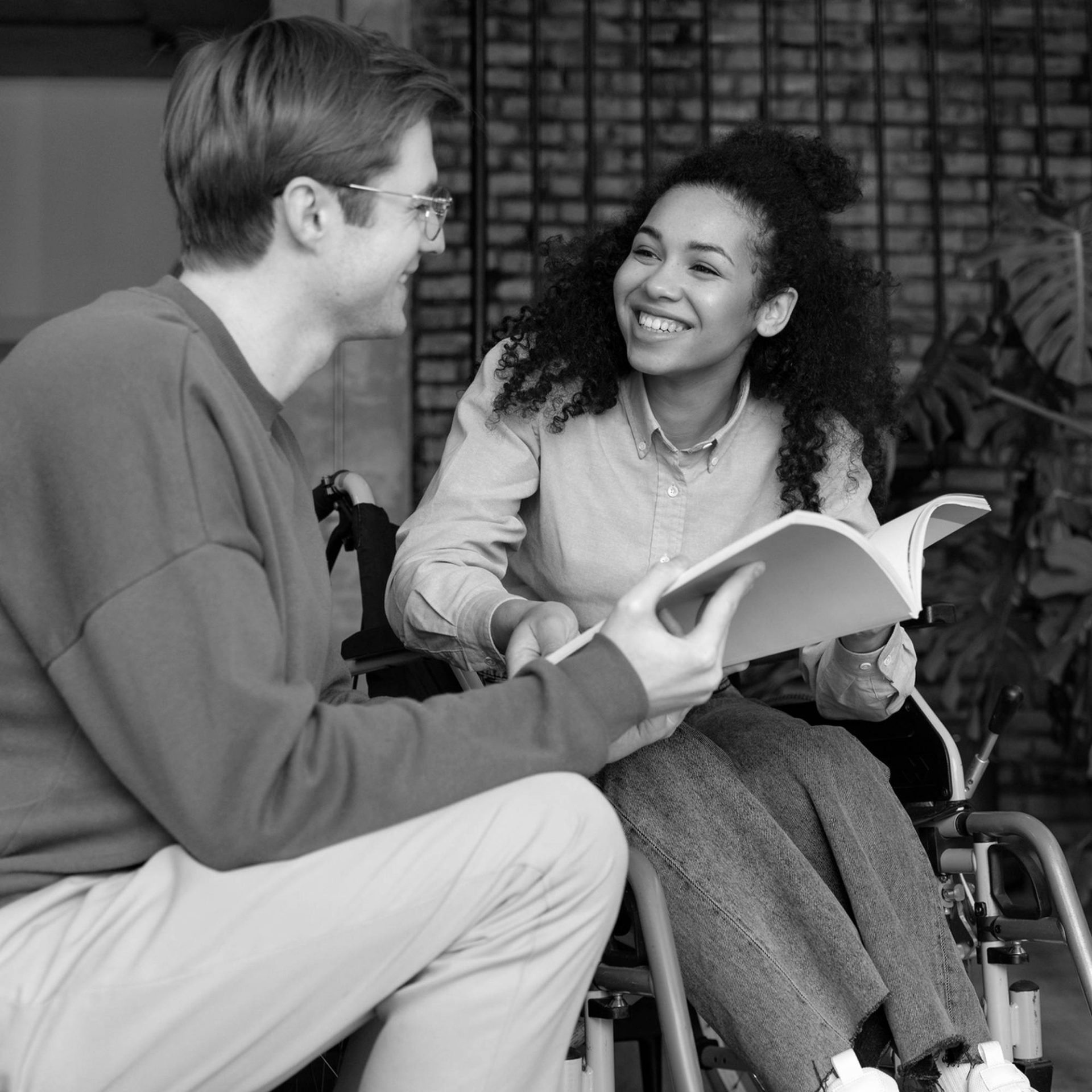 a person in a wheel chair shows another person a page in a book as they smile at each other