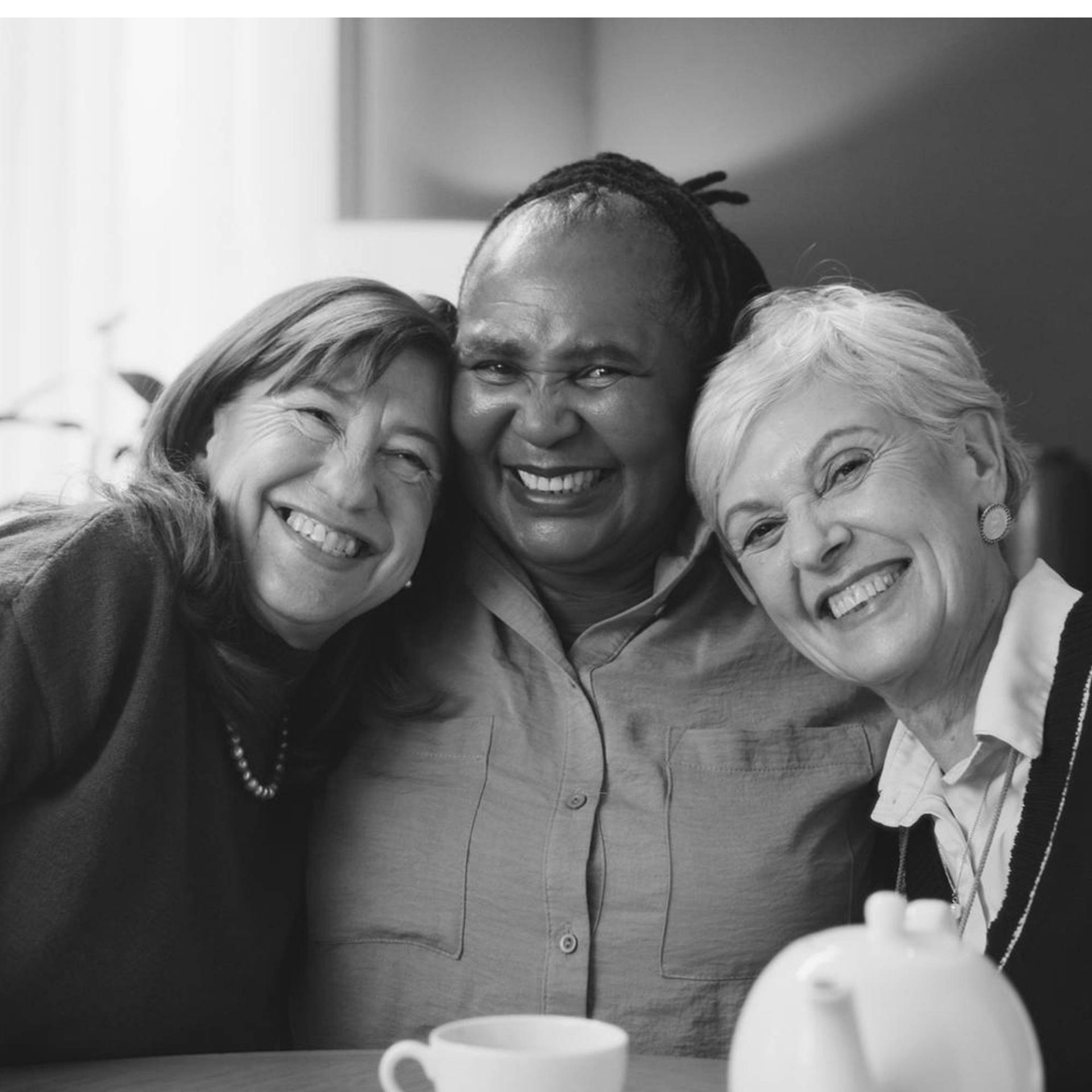 three diverse, older women hug each other and smile at the viewer